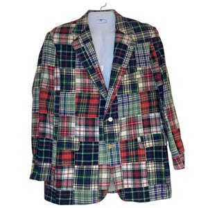 May include: A patchwork blazer in a variety of plaid patterns, including red, green, navy, and white. The jacket has a classic two-button closure and is hanging on a white hanger. The blazer is a mix of different plaid fabrics.