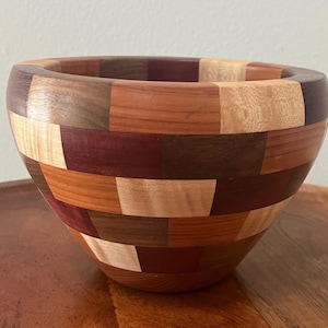 May include: A handcrafted wooden bowl with a mosaic design. The bowl is constructed from various wood types, featuring a patchwork pattern of light and dark brown, red, and tan rectangular pieces. The bowl has a rounded shape and a smooth finish.