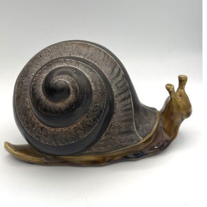 May include: A ceramic snail figurine featuring a dark brown, spiraled shell with fine, etched linear textures. The body of the snail is a smooth, glossy, light brown glaze that extends from the head to the tail, showcasing a contrast in finish.