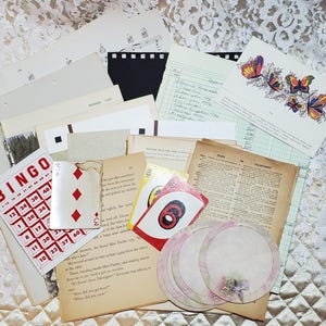 Puede incluir: Una colección de efímeras de papel vintage, que incluye una tarjeta de bingo, cartas de juego, una página de un libro, una hoja de música y un libro mayor. Los papeles están dispuestos sobre un mantel de encaje blanco.