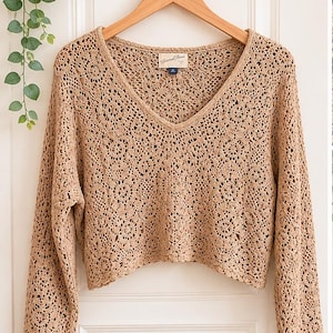 May include: A tan, long-sleeved crochet top with a V-neck. The top is hanging on a wooden hanger against a white door. The crochet pattern is intricate and open, with a scalloped hemline. The tag reads "Universal Thread".