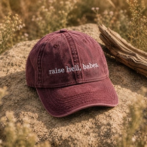 以下が含まれることがあります： 「raise hell, babes」の刺繍が入ったマルーン色のベースボールキャップ。柔らかく使い古された素材でできており、つばがカーブしています。帽子は岩の上に置かれています。