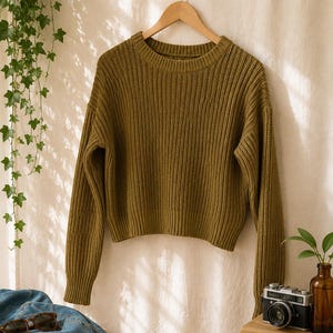 Könnte beinhalten: Ein olivgrüner Langarmpullover mit gerippter Strickstruktur, Rundhalsausschnitt sowie gerippten Bündchen an Ärmeln und Saum. Der Pullover hängt an einem Holzkleiderbügel vor einem neutralen Stoffhintergrund und zeigt ein schlichtes Design.