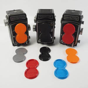 Puede incluir: Tres cámaras réflex de doble objetivo vintage, dos etiquetadas como Rolleicord y una Fodorflex, están dispuestas con varias tapas de lente en forma de ocho en naranja, negro, rojo, gris y azul. Las tapas presentan texto en relieve.