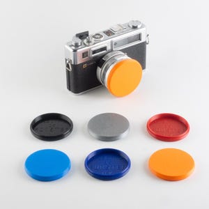 Pode incluir: Uma câmara vintage prateada e preta com uma tampa de lente laranja. Abaixo estão seis tampas de lente circulares adicionais em preto, prata, vermelho, azul, azul escuro e laranja. Cada tampa apresenta o texto em relevo FORSTER UK.