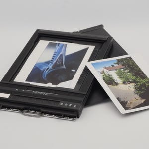 Op de afbeelding: Een zwarte filmhouder voor grootformaat fotografie met het woord Riteway op het frame. Een vierkante instantfoto met een blauw mechanisch detail zit in de houder, en een tweede rechthoekige foto van een gebouw ligt ernaast.
