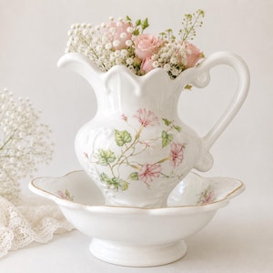 May include: A white porcelain pitcher and basin set, adorned with a floral design of pink flowers and green leaves. The pitcher is filled with pink roses and baby's breath. The basin has a gold trim.