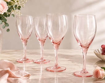 May include: Four pink-tinted wine glasses with long stems are arranged on a marble surface. A vase of pink roses and a small bowl of grapes are in the background. A pink cloth is draped on the left.