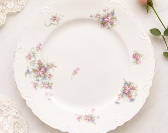 May include: White porcelain plate with a scalloped edge, adorned with delicate pink and blue floral clusters. The plate has a gold trim and is set against a white background, evoking a vintage aesthetic. A pink rose is visible in the background.
