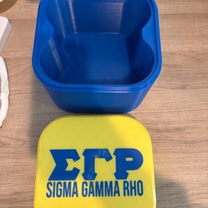Op de afbeelding: Een blauwe, 3D-geprinte container met een vierkante vorm en afgeronde randen. Een gele deksel met de tekst "ΣΓΡ SIGMA GAMMA RHO" in blauw staat naast de container. De container heeft twee gebogen inkepingen aan de zijkanten.