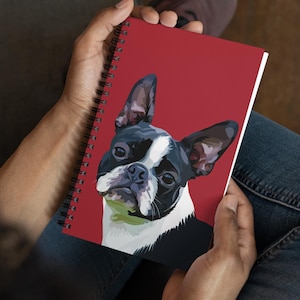 May include: A spiral-bound notebook featuring a Boston Terrier dog illustration. The dog is depicted in black, white, and brown against a red background. The notebook is held open, showcasing the dog's face and ears.