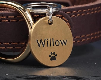 May include: A round brass pet ID tag engraved with the name Willow and a paw print icon, attached to a silver-toned split ring. The tag is suspended from a dark brown leather collar featuring visible stitching and a polished gold-toned metal D-ring.