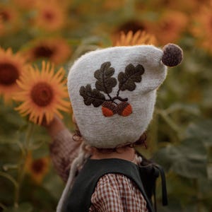 以下が含まれることがあります： 刺繍されたオークの葉とドングリが付いた、茶色のポンポンが付いたライトグレーのニット帽。 ひまわり畑で、濃い緑色のドレスと赤と白のチェック柄のシャツを着た人がかぶっています。