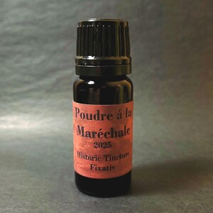 May include: A small, dark glass bottle with a black cap. The bottle has a red label with the text "Poudre à la Maréchale 2025 Historic Tincture Fixativ". The bottle is likely a historic tincture.