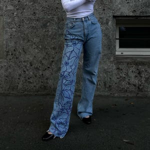 Pode incluir: Jeans de denim azul com um design floral em uma perna. O design é de cor azul escura e apresenta um padrão repetitivo de flores.