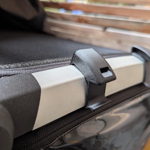 Peut inclure: Un clip en plastique noir fixé sur le cadre en métal argenté d'une poussette. Le clip présente une surface texturée et une petite ouverture rectangulaire. Le cadre est positionné contre une capote en tissu noir et une fenêtre en plastique.