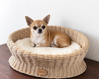 May include: An oval-shaped light brown woven wicker pet bed featuring a soft cream-colored plush cushion interior. An oval wooden name tag with the engraved name Penny is attached to the front of the wicker frame, which holds a small tan dog.