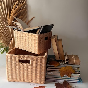 May include: Two woven wicker baskets with handles, one stacked on top of the other. The top basket is filled with books. A stack of books with autumn leaves sits next to the baskets.