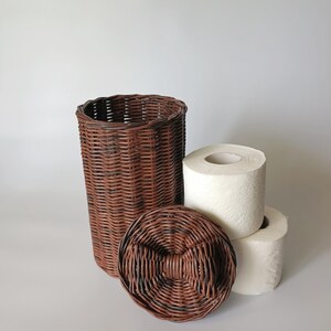 Brown toilet paper storage basket with a close-up of the lid featuring a woven bow; two rolls beside for reference. Cozy and unique bathroom piece.