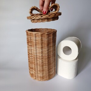 Natural round toilet paper container with a hand lifting the lid; two rolls beside it. Practical and decorative solution.