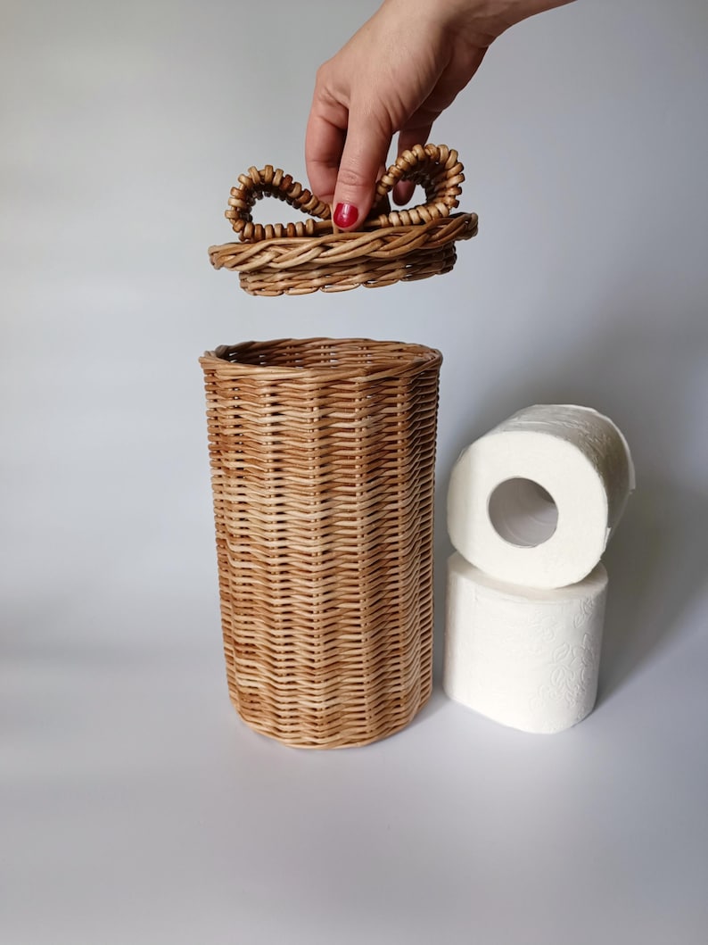 Natural round toilet paper container with a hand lifting the lid; two rolls beside it. Practical and decorative solution.
