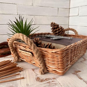May include: A woven wicker basket with rope handles and a wooden tag that says "Hand Made". The basket is filled with a small potted plant, pine cones, and a book.