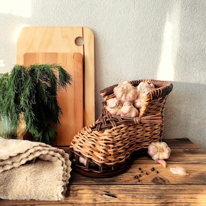 May include: A brown woven wicker basket shaped like a shoe, filled with several white garlic bulbs. The basket sits on a wooden surface next to a bunch of fresh green dill, two wooden cutting boards, and a folded beige textured cloth.