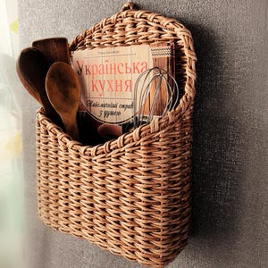 Könnte beinhalten: Ein an der Wand montierter, geflochtener Weidenkorb mit zwei Holzlöffeln, einem Metallschneebesen und einem Buch mit dem Titel "Українська кухня". Der Korb hat eine abgerundete Oberseite mit einer Schlaufe und einen rechteckigen Korpus.