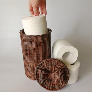 Hand pulling a roll of toilet paper from the brown wicker basket; lid with bow placed nearby. Shows functionality and size.