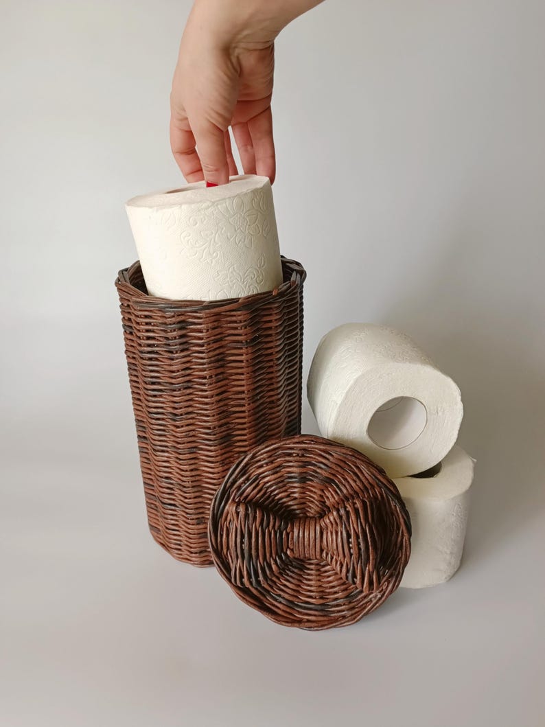 Hand pulling a roll of toilet paper from the brown wicker basket; lid with bow placed nearby. Shows functionality and size.