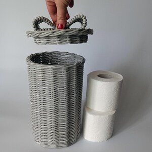 Gray woven toilet roll bin with a hand holding the lid above it; paper rolls beside it. Handmade rattan-style bathroom basket.
