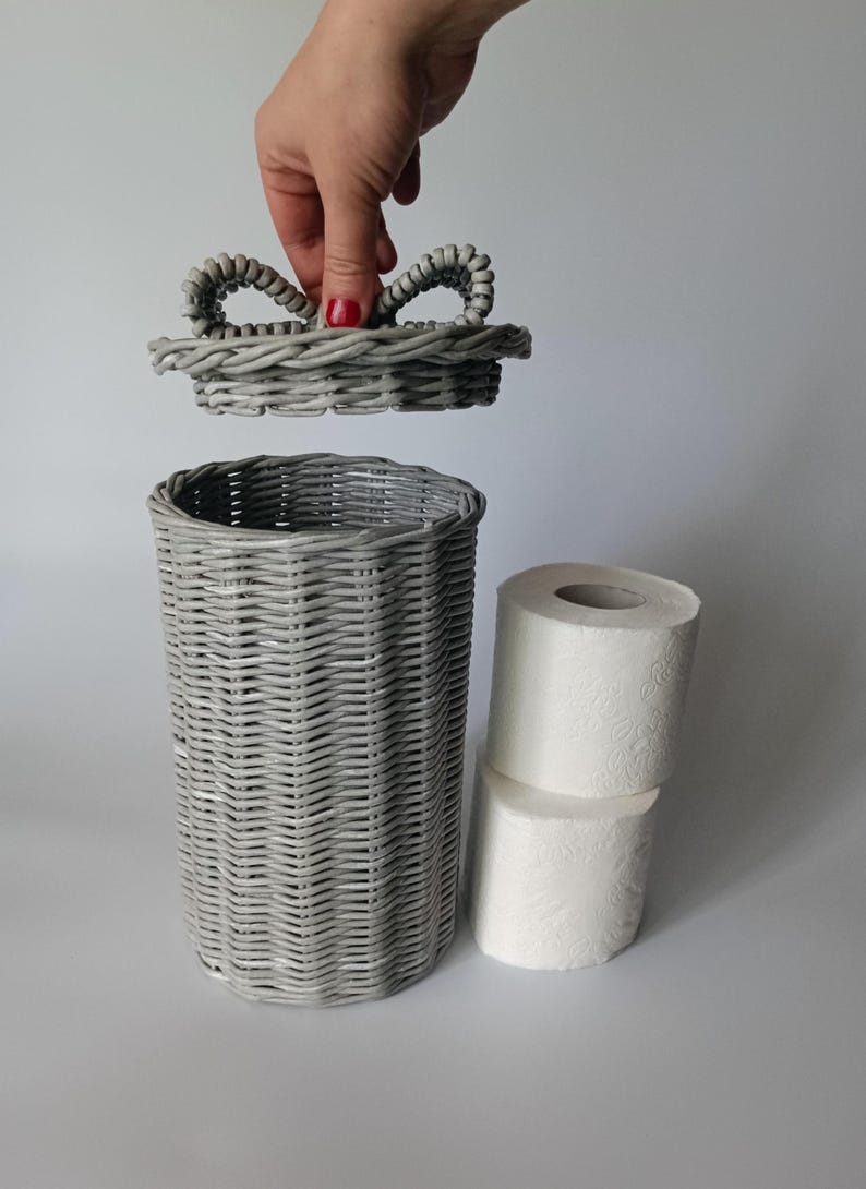 Gray woven toilet roll bin with a hand holding the lid above it; paper rolls beside it. Handmade rattan-style bathroom basket.