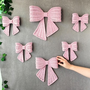 May include: A collection of six light pink woven wicker bows in varying sizes. Each bow features a central knot and two downward-hanging tails with a textured, interlaced pattern. The bows are arranged in a cluster, showcasing a consistent light pink color and a uniform woven construction throughout the set.