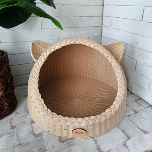 May include: A light beige woven pet bed with a rounded dome shape and two pointed cat-like ears on top. The front opening features a braided trim, and a small oval wooden plaque with the engraved text Mew is attached to the lower front edge.