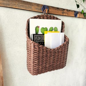 May include: A brown woven wall-mounted basket organiser featuring a curved top handle. The basket holds a white card with a green cactus graphic, a black notebook with a white paisley pattern, and a plain white envelope tucked inside the pocket.