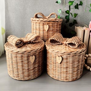 May include: Three woven wicker baskets in varying sizes. Each basket is round with a lid and a bow-shaped handle. A small wooden heart is attached to the front of each basket. The baskets are light brown in color.