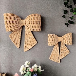 May include: Two wall-mounted bow decorations crafted from light tan woven wicker. The larger bow is positioned on the left, while a smaller, matching bow is located to the right. Both items feature a classic bow shape with two long, hanging tails.