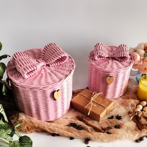 May include: Two pink woven baskets with bow-shaped lids and heart-shaped wooden accents. A small wrapped gift and decorative items are also present. The baskets are round and have a light pink color, suitable for storage or decoration.
