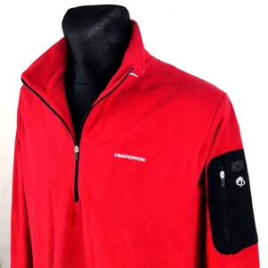 Peut inclure: Une veste polaire rouge avec une fermeture éclair noire et une poche noire sur la manche gauche. La veste porte le nom de la marque "CRAGHOPPERS" en lettres blanches sur la poitrine. La veste est présentée sur un mannequin noir.
