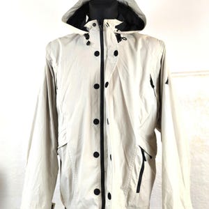 May include: A beige hooded jacket with a zip and snap fastening. The jacket has black accents and two zipped pockets on the front.