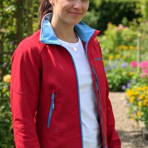 May include: A red jacket with a light blue collar and zipper trim. The jacket is zipped up and has a white t-shirt underneath. The background is a garden with green foliage and colorful flowers.