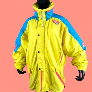 Peut inclure: Une combinaison de ski jaune vintage avec des accents bleus et roses. La veste a un col montant, des boutons et un cordon de serrage. Les manches ont des panneaux bleus, et la combinaison comporte des fermetures éclair roses et un écusson. Une combinaison de ski rétro.