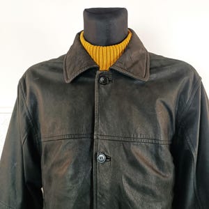 Op de afbeelding: Een zwart leren jack met een knoopsluiting en een kraag. Het jack heeft een licht verweerde look en wordt gedragen op een mannequin.