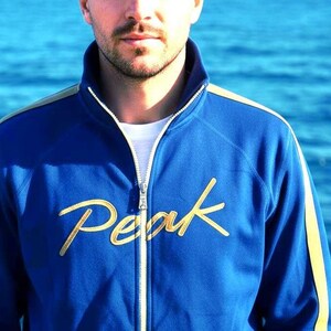 May include: A blue zip-up jacket with gold detailing and the word "Peak" embroidered on the front. The jacket features a white zip and a white undershirt. The background is a blurred view of the sea.