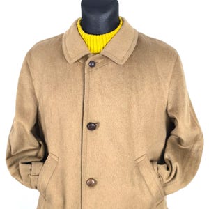 May include: A tan wool coat with a classic collar and three dark brown buttons. The coat has two front pockets and is worn over a bright yellow turtleneck sweater. The coat is displayed on a black mannequin head.