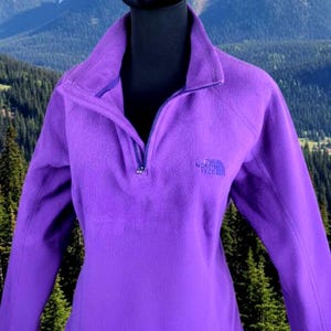 Op de afbeelding: Een paarse fleece trui met een halve ritssluiting en een opstaande kraag. Het The North Face-logo is zichtbaar op de borst. De achtergrond toont een berglandschap.