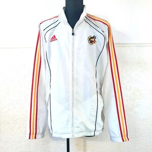 May include: White Adidas track jacket with red, yellow, and black stripes on the sleeves. The jacket has a zipper closure and a small Adidas logo on the left chest. The Spanish national team crest is on the right chest.