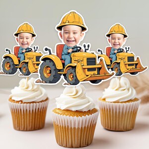 Puede incluir: Tres adornos para cupcakes de vehículos de construcción amarillos y negros con caras de dibujos animados. Cada adorno presenta a una persona con un sombrero de construcción amarillo conduciendo un tractor amarillo con una pala negra.