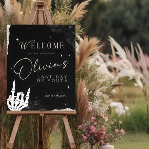 May include: Black and white welcome sign with a skeleton hand making a rock and roll gesture. The sign reads "Welcome, We are mourning Olivia's Last Day of Youth, XIII-IX-MMXXV".