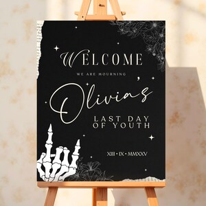 May include: Black welcome sign with white text that reads "Welcome, We are mourning Olivia's Last Day of Youth, XIII - IX - MMXXV". The sign has a white skeleton hand giving the middle finger and floral designs.